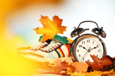 Photo of Alarm clock, dry leaves and stylish sneakers on blurred background, closeup