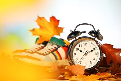 Photo of Alarm clock, dry leaves and stylish sneakers on blurred background, closeup