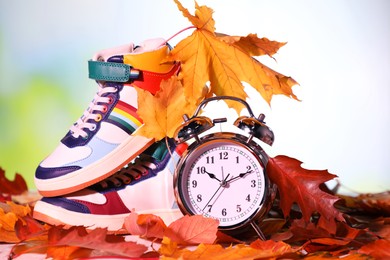 Photo of Alarm clock, dry leaves and stylish sneakers on blurred background, closeup