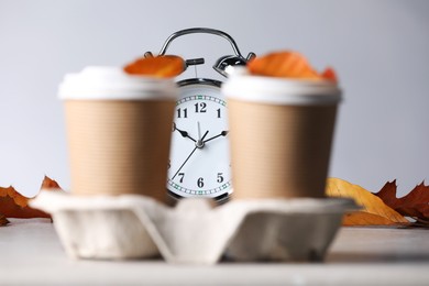 Photo of Alarm clock, dry leaves and cups of coffee on light grey background, closeup