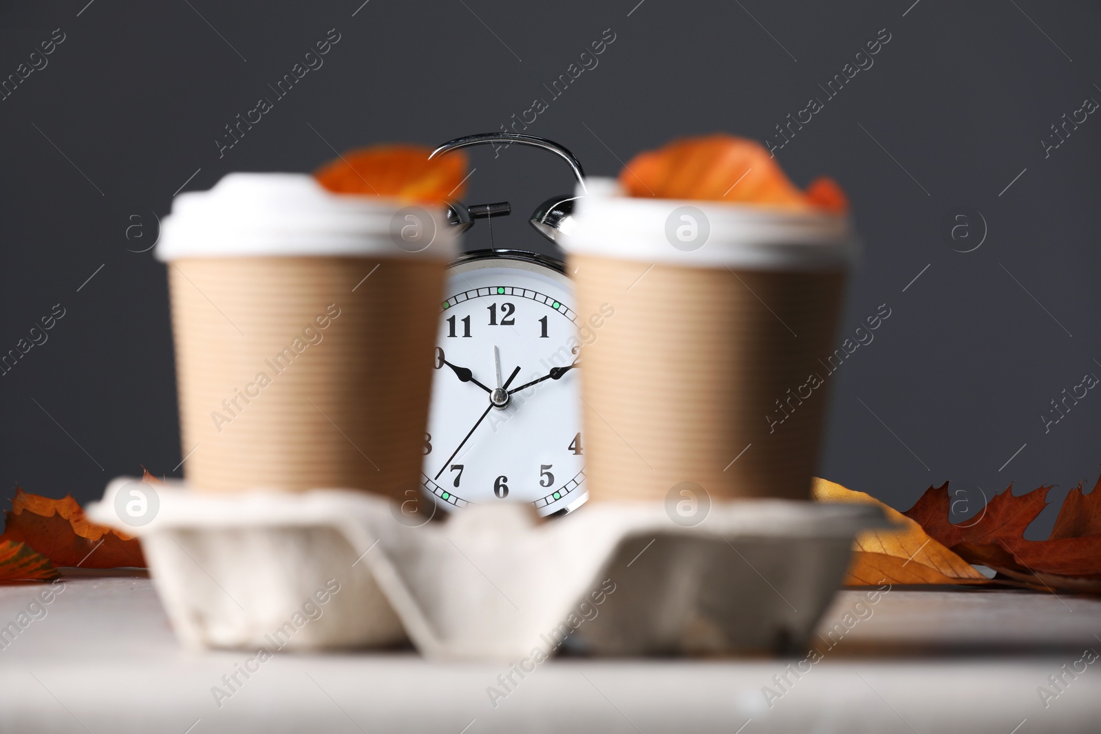 Photo of Alarm clock, dry leaves and cups of coffee on light grey background, closeup