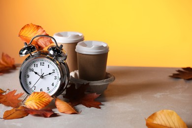 Photo of Alarm clock, dry leaves and cups of coffee on light grey table against yellow background, space for text