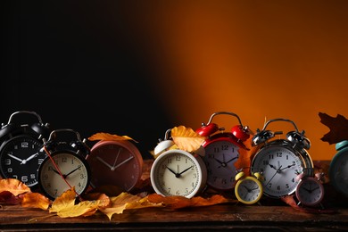 Photo of Alarm clocks and dry leaves on wooden table against brown background, space for text