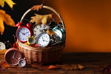 Photo of Alarm clocks and dry leaves in wicker basket on wooden table against brown background, space for text