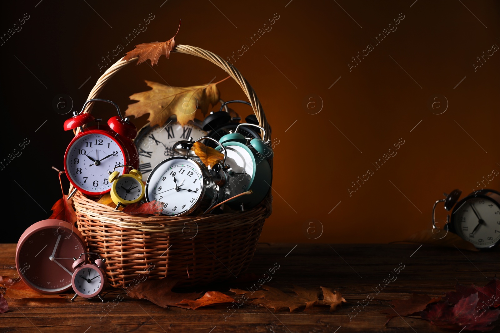 Photo of Alarm clocks and dry leaves in wicker basket on wooden table against brown background, space for text