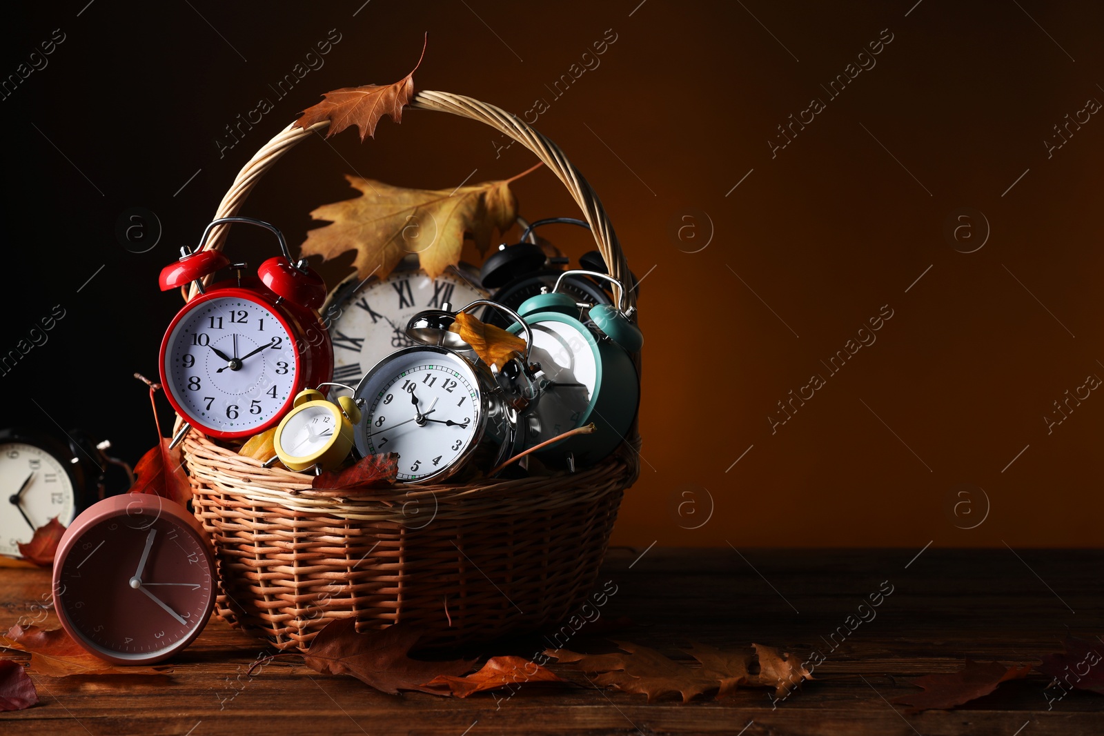 Photo of Alarm clocks and dry leaves in wicker basket on wooden table against brown background, space for text