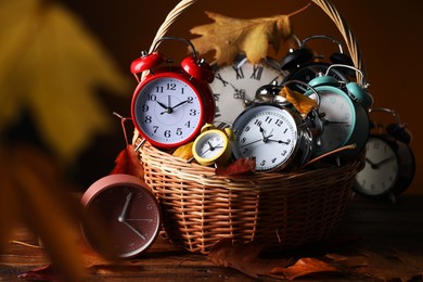 Photo of Alarm clocks and dry leaves in wicker basket on wooden table against brown background