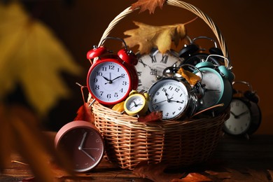 Photo of Alarm clocks and dry leaves in wicker basket on wooden table against brown background