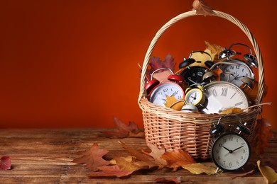 Photo of Alarm clocks and dry leaves in wicker basket on wooden table against brown background, space for text