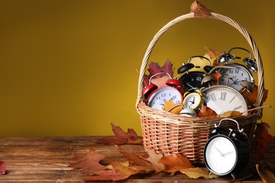 Photo of Alarm clocks and dry leaves in wicker basket on wooden table against olive background, space for text