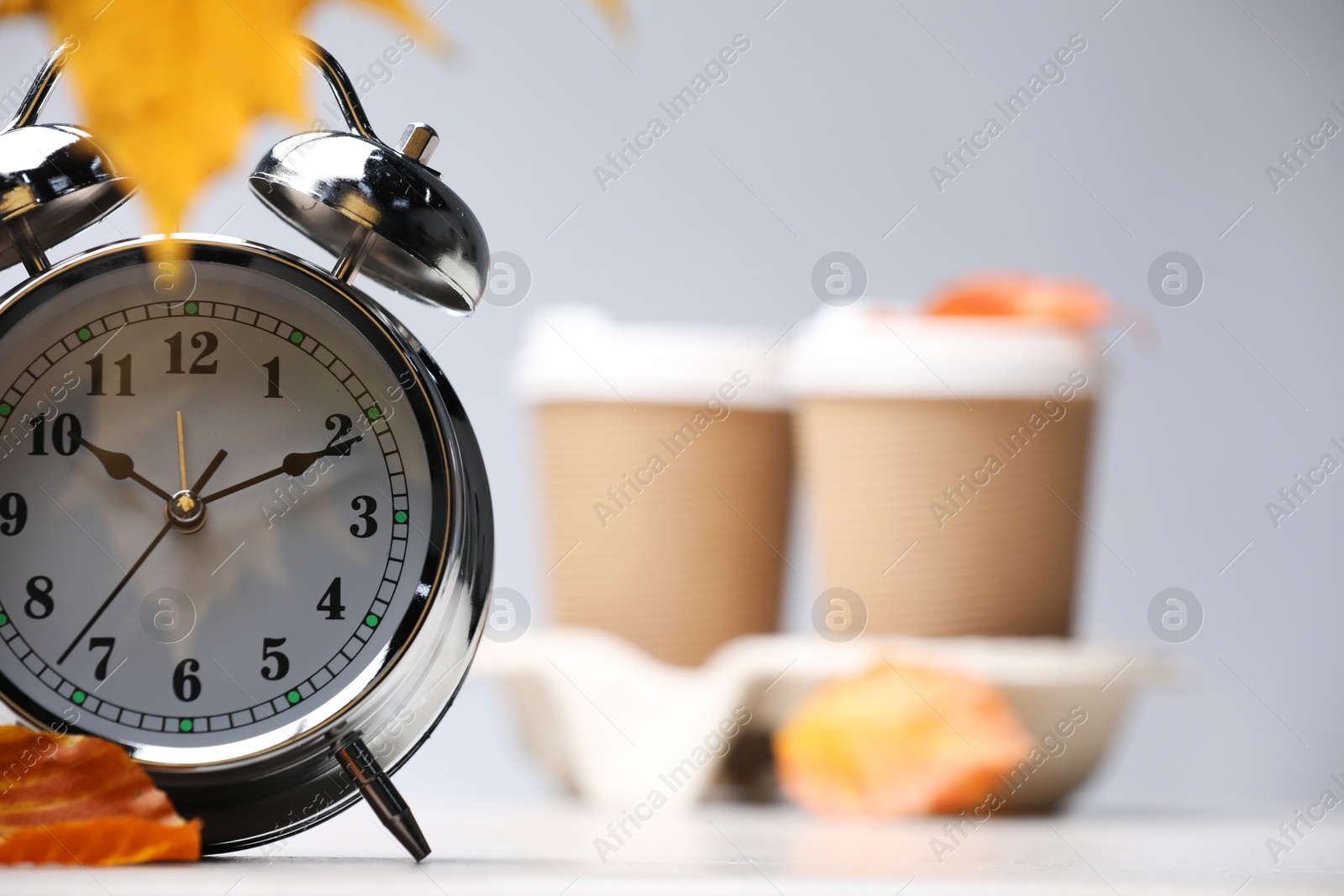 Photo of Alarm clock, dry leaves and cups of coffee on light grey background, closeup. Space for text