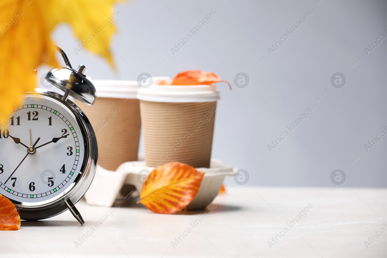 Photo of Alarm clock, dry leaves and cups of coffee on light grey background, closeup. Space for text