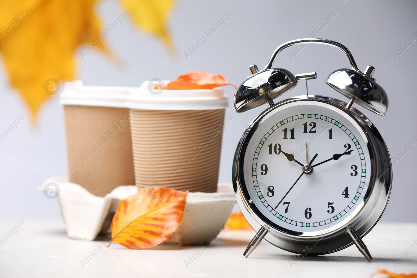 Photo of Alarm clock, dry leaves and cups of coffee on light grey background, closeup