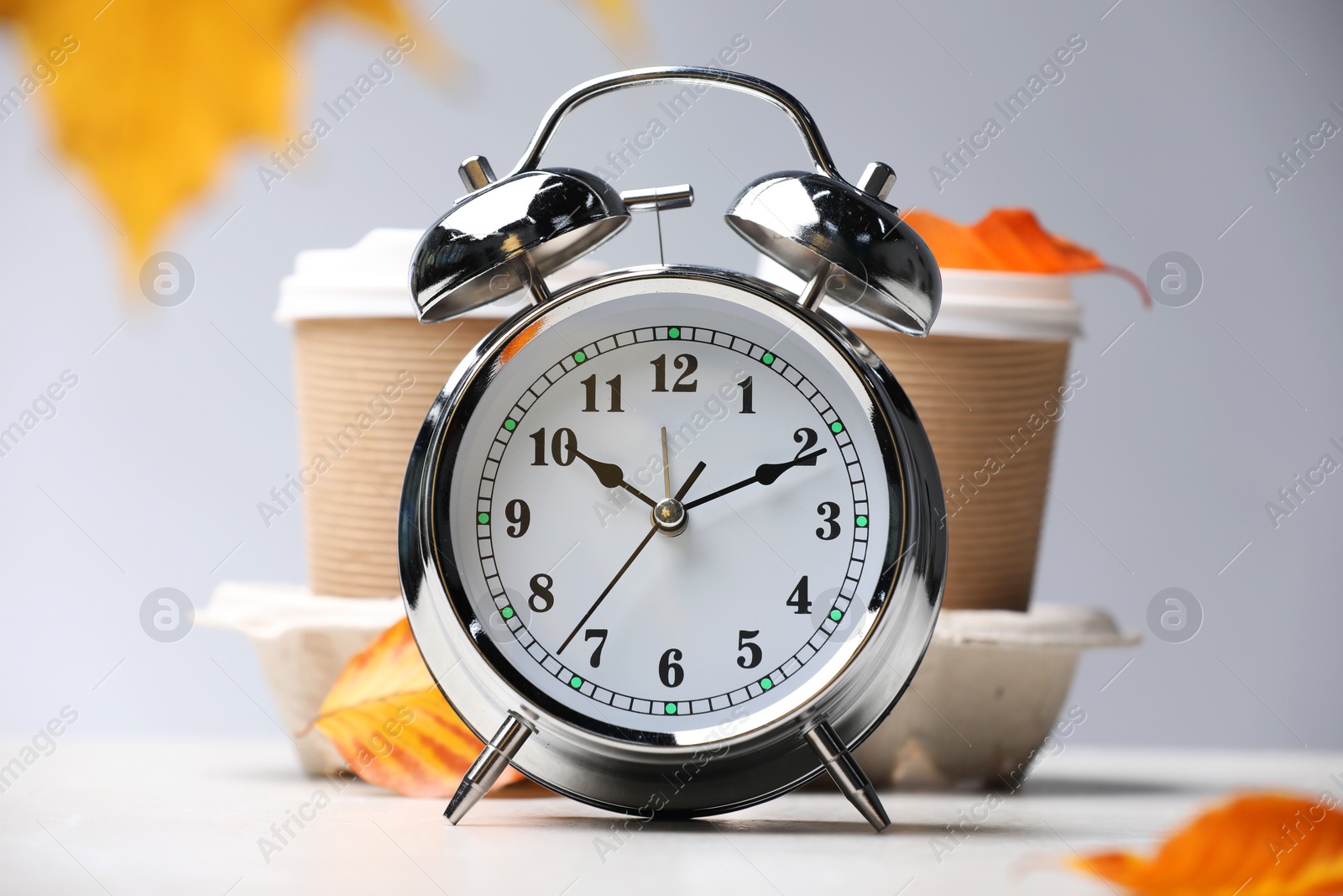 Photo of Alarm clock, dry leaves and cups of coffee on light grey background, closeup