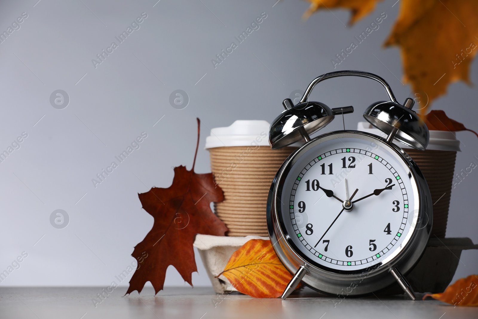 Photo of Alarm clock, dry leaves and cups of coffee on light grey background, closeup. Space for text