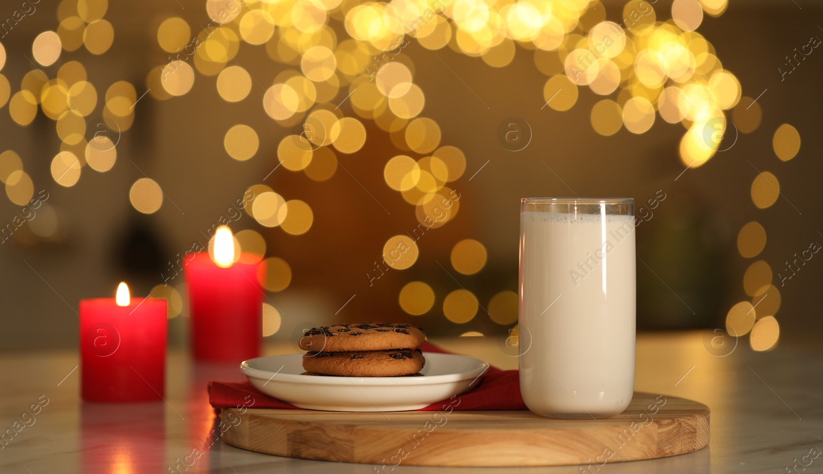 Photo of Glass of milk, tasty chocolate chip cookies and burning candles on table against blurred Christmas lights. Bokeh effect