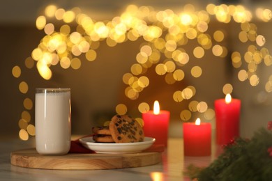 Photo of Glass of milk, tasty chocolate chip cookies and burning candles on table against blurred Christmas lights. Bokeh effect