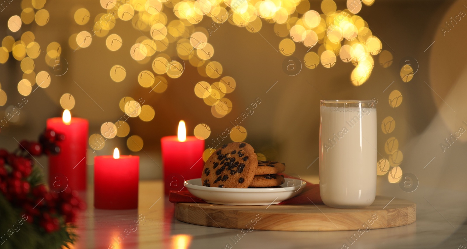 Photo of Glass of milk, tasty chocolate chip cookies and burning candles on table against blurred Christmas lights. Bokeh effect