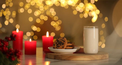 Photo of Glass of milk, tasty chocolate chip cookies and burning candles on table against blurred Christmas lights. Bokeh effect