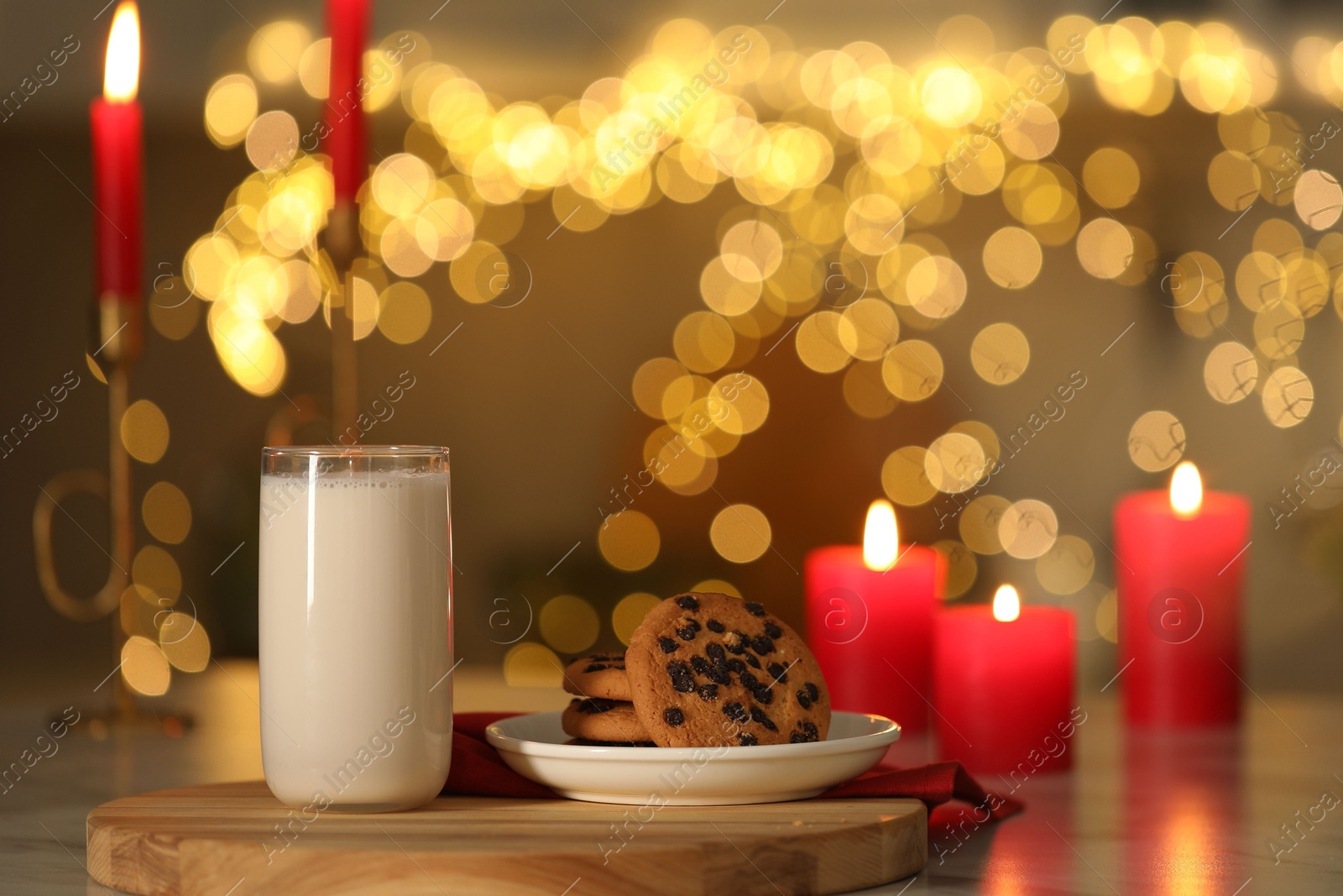 Photo of Glass of milk, tasty chocolate chip cookies and burning candles on table against blurred Christmas lights. Bokeh effect