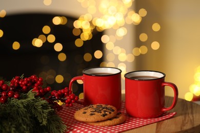 Photo of Tasty cocoa in cups, chocolate chip cookies and decorative branches on table against blurred Christmas lights. Bokeh effect