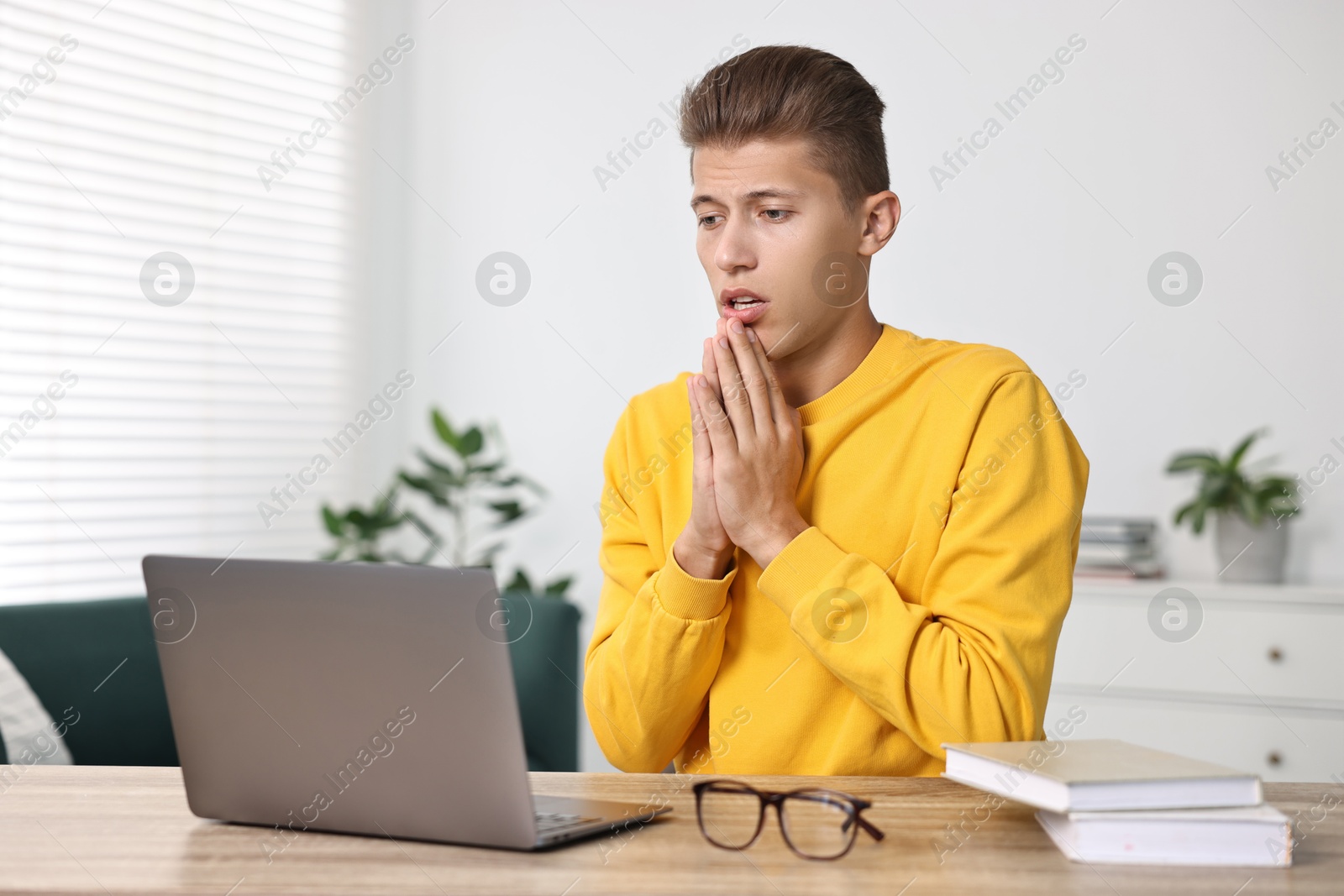 Photo of Worried student praying for good exam result at table with laptop indoors