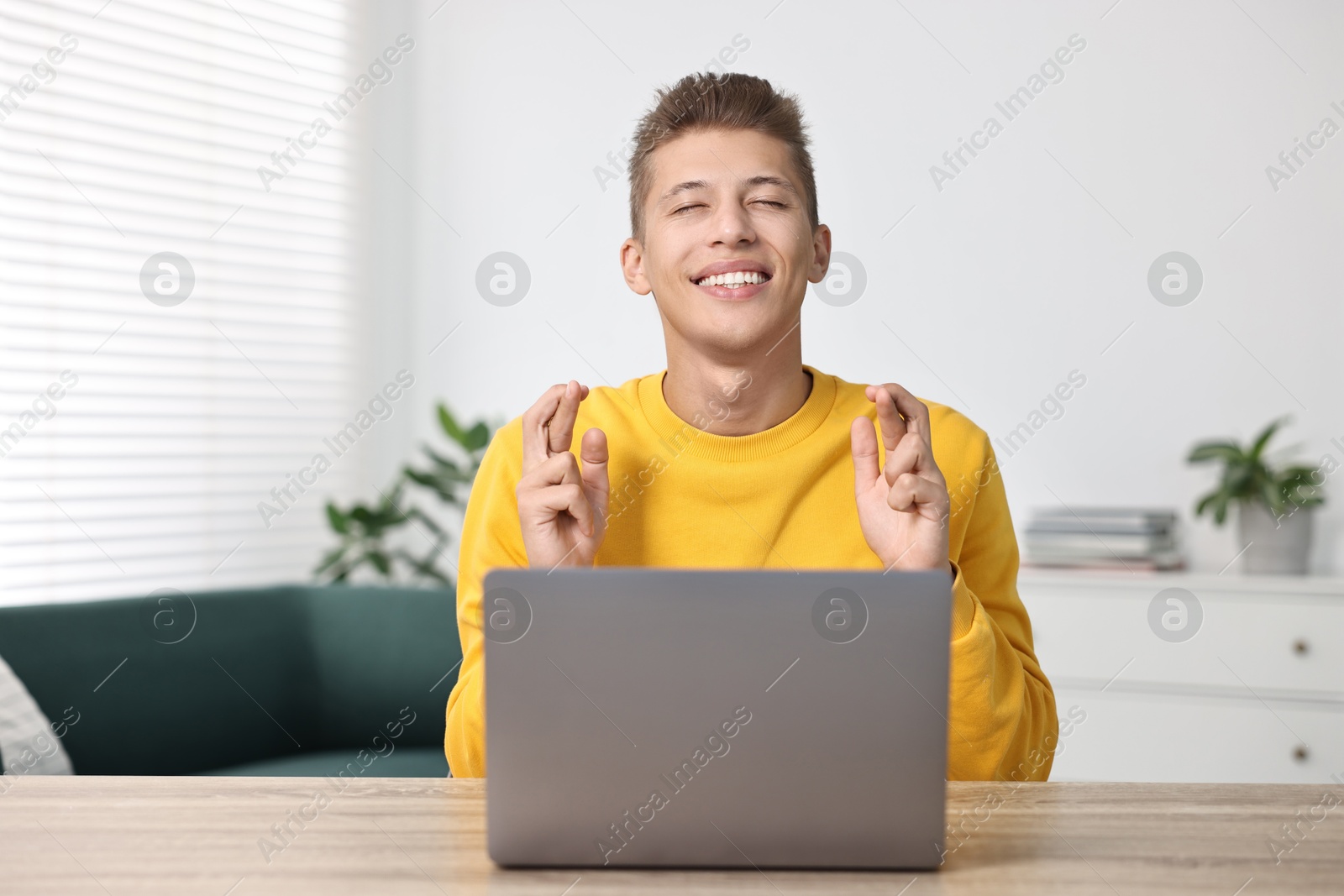 Photo of Student with crossed fingers at table with laptop indoors. Hope for good exam result