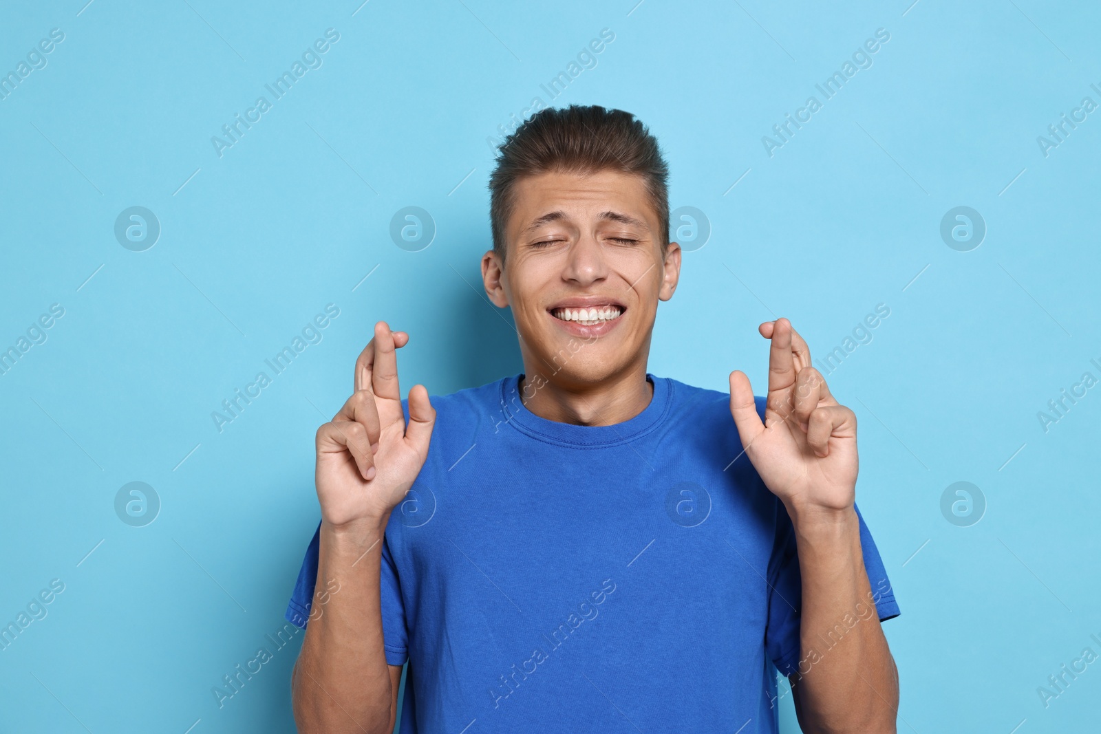 Photo of Emotional student with crossed fingers on light blue background. Hope for good exam result