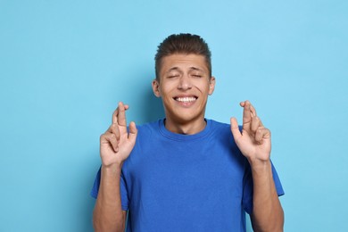 Photo of Emotional student with crossed fingers on light blue background. Hope for good exam result