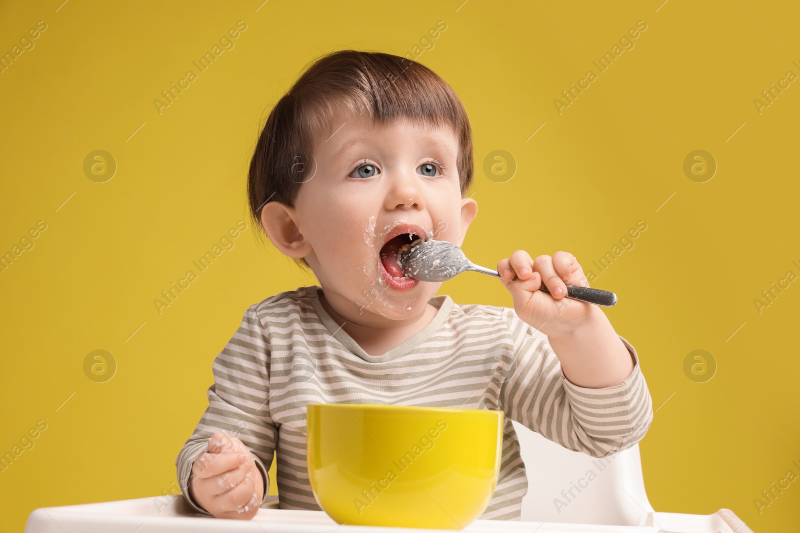 Photo of Cute little kid eating healthy baby food from bowl in high chair on yellow background