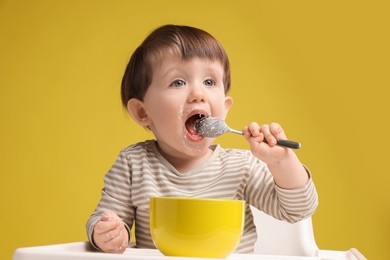 Photo of Cute little kid eating healthy baby food from bowl in high chair on yellow background