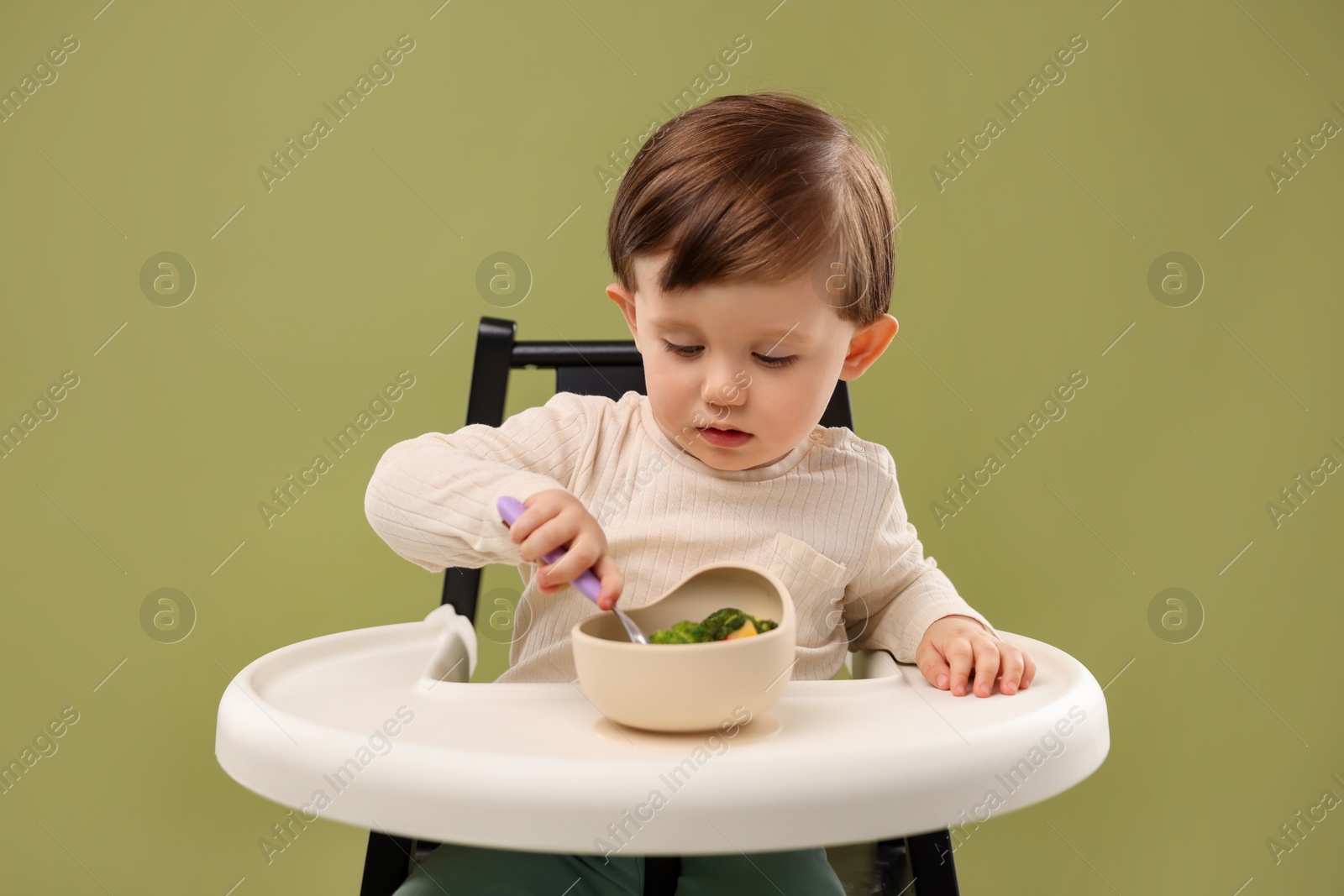 Photo of Cute little baby eating healthy food from bowl in high chair on olive background