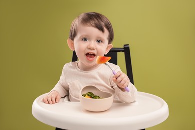 Photo of Cute little baby eating healthy food from bowl in high chair on olive background