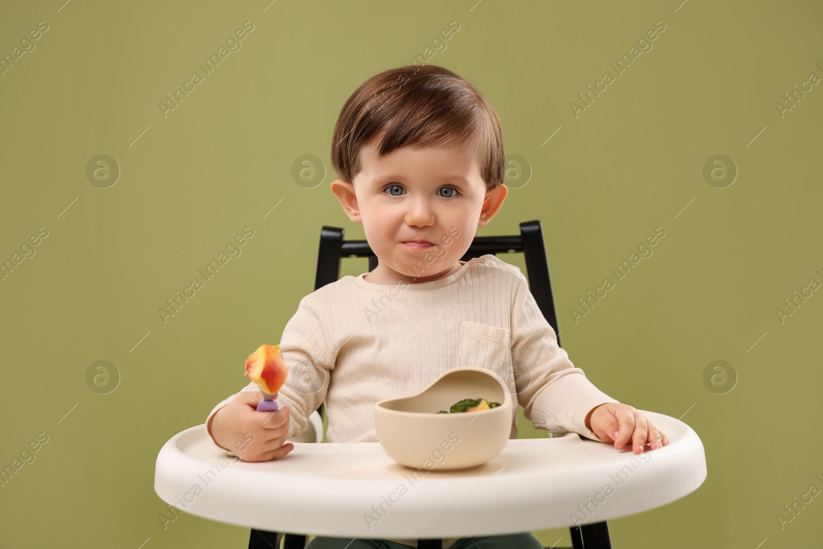 Photo of Cute little baby eating healthy food from bowl in high chair on olive background