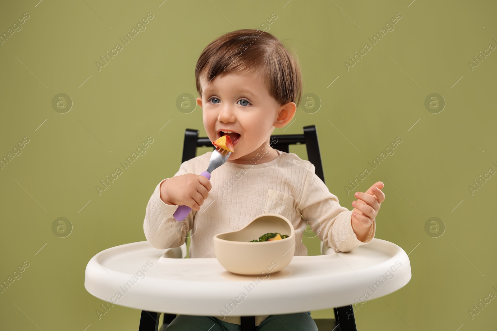 Photo of Cute little baby eating healthy food from bowl in high chair on olive background