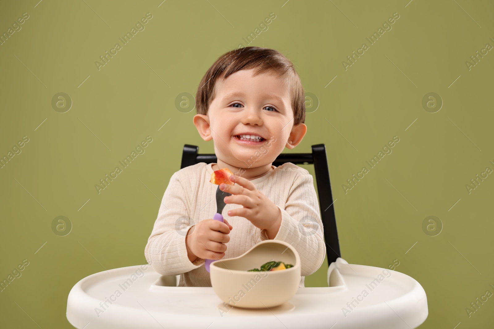 Photo of Cute little baby eating healthy food from bowl in high chair on olive background