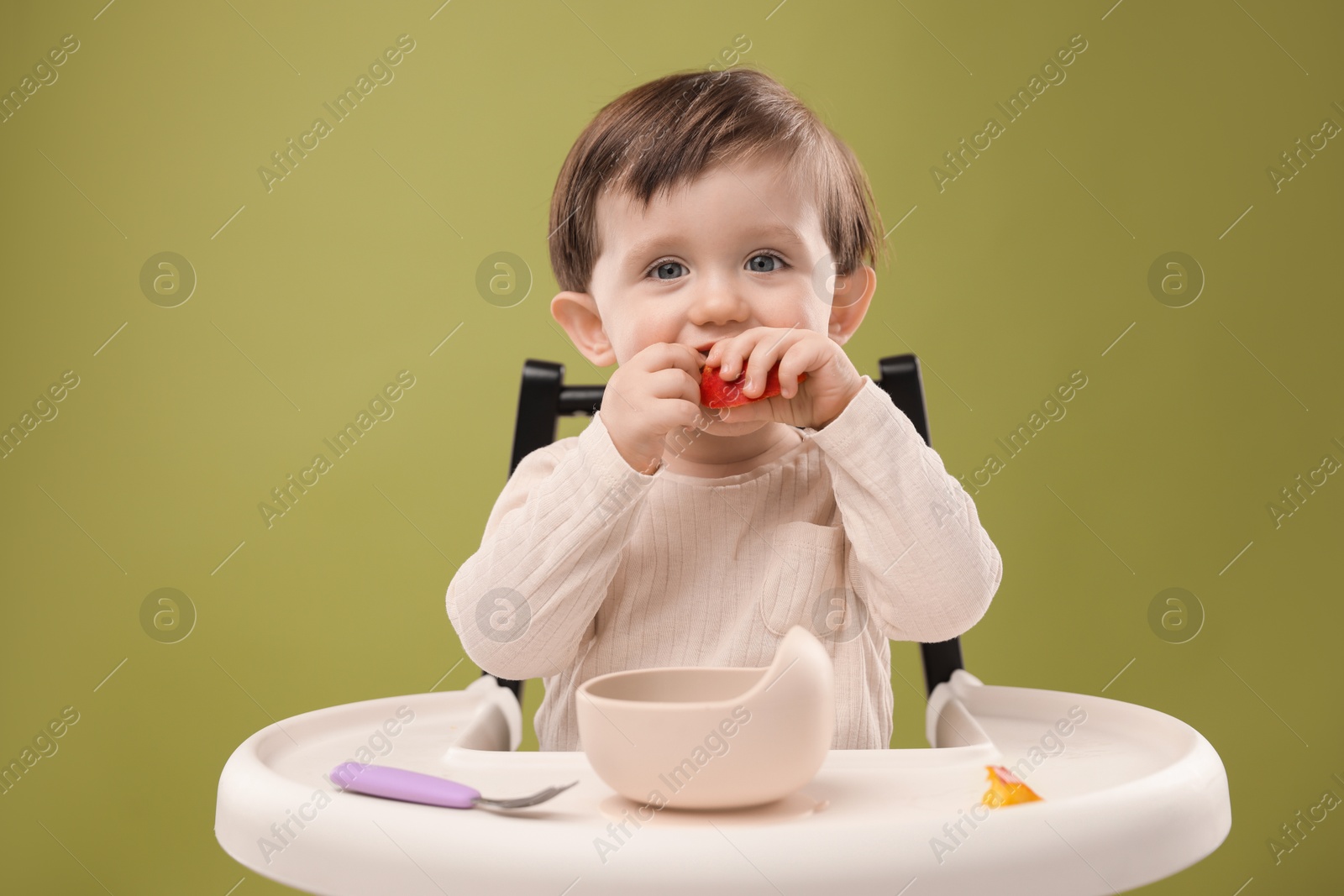 Photo of Cute little baby eating healthy food from bowl in high chair on olive background