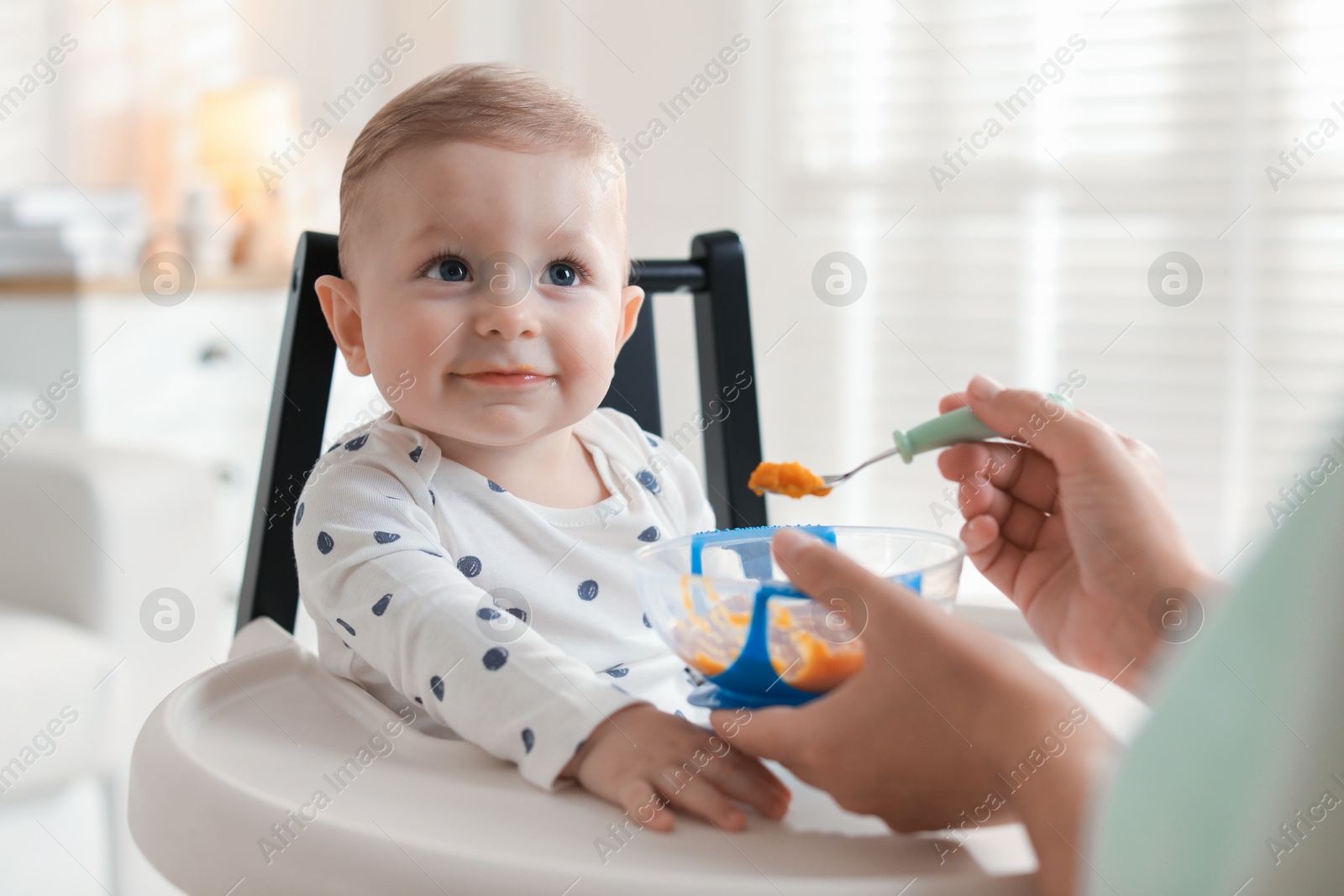 Photo of Mother feeding her cute little baby in high chair at home, closeup