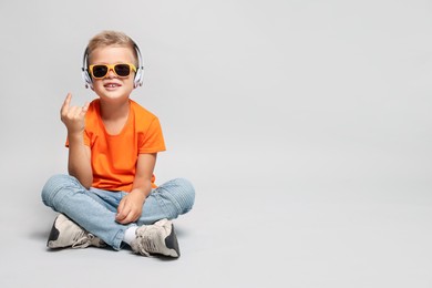 Little boy with sunglasses listening to music and showing rock gesture on light grey background, space for text