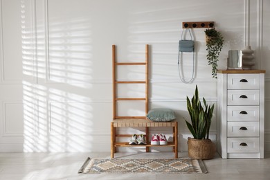 Photo of Stylish hallway interior with shoe storage bench, chest of drawers, houseplants and coat rack