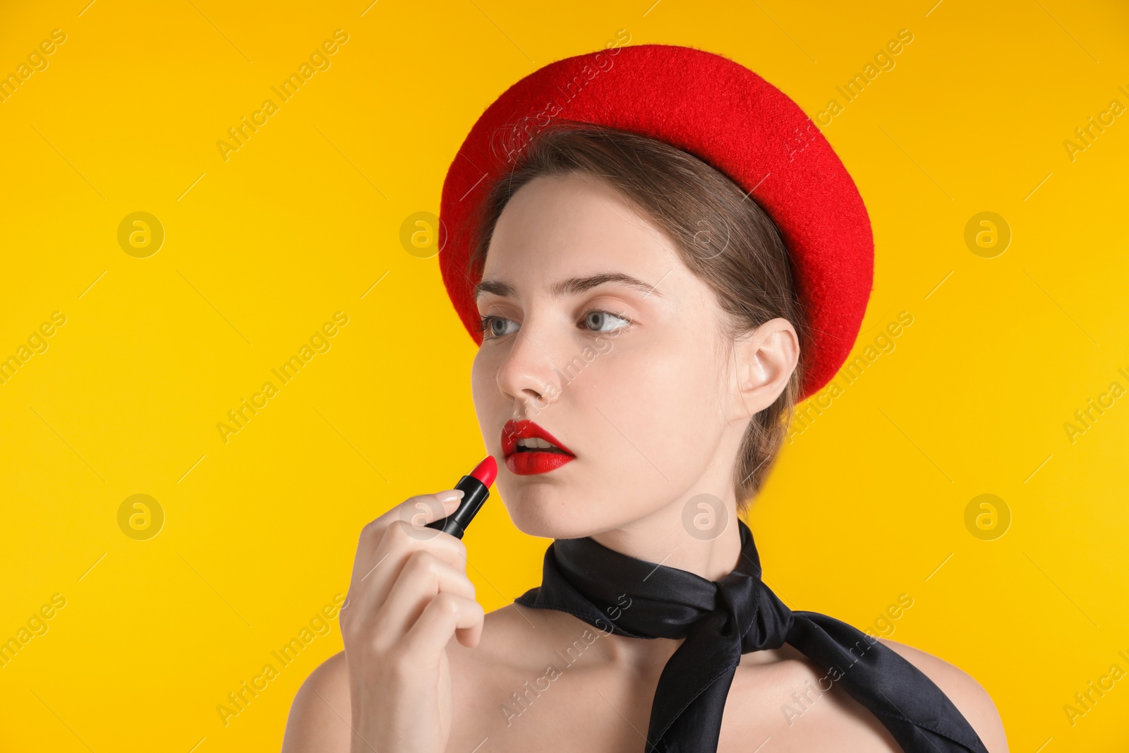 Photo of Beautiful young woman with red beret and black neckerchief applying lipstick against yellow background