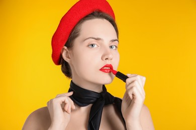 Photo of Beautiful young woman with red beret and black neckerchief applying lipstick against yellow background