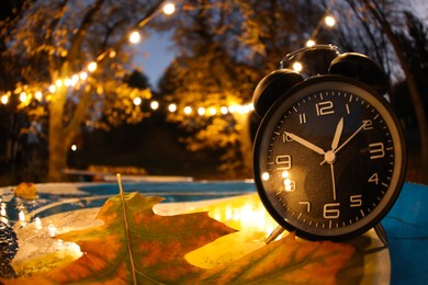 Photo of Autumn time. Alarm clock and fallen leaves on blue surface in evening park, wide angle lens