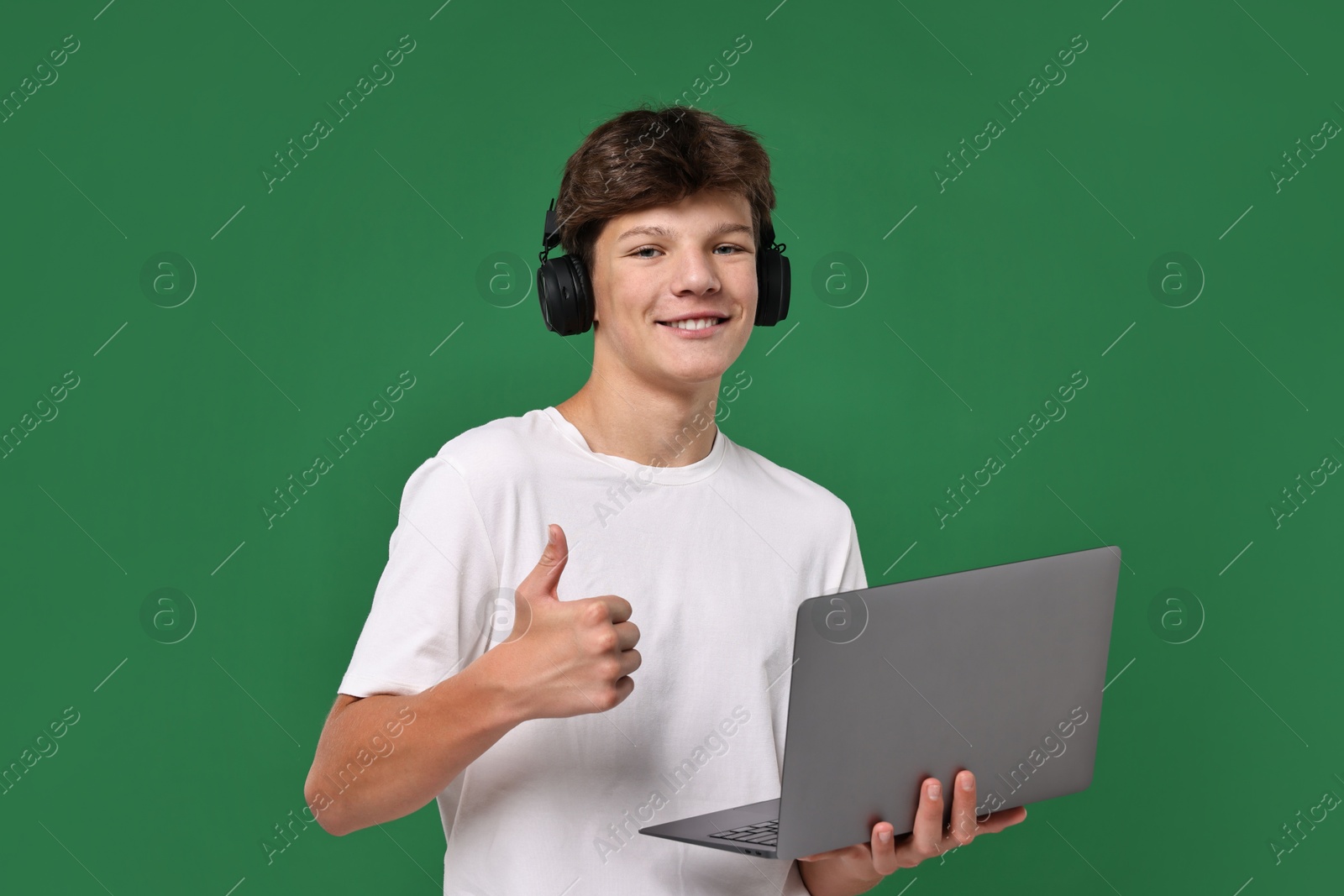 Photo of Teenage boy in headphones with laptop showing thumbs up on green background