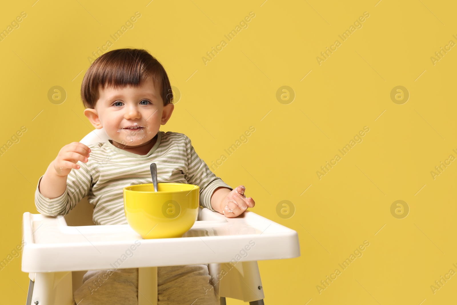 Photo of Cute little kid eating healthy baby food from bowl in high chair on yellow background, space for text