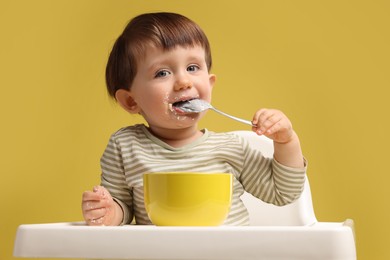 Photo of Cute little kid eating healthy baby food from bowl in high chair on yellow background