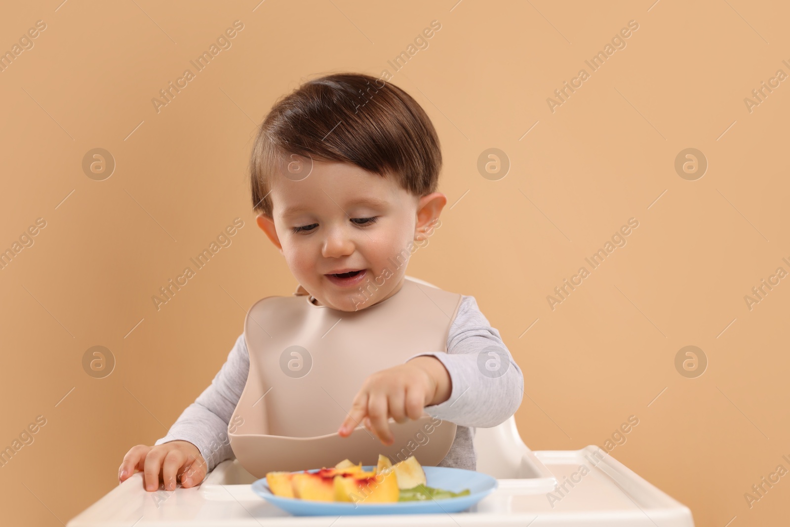 Photo of Healthy baby food. Cute little kid eating fruits in high chair on beige background