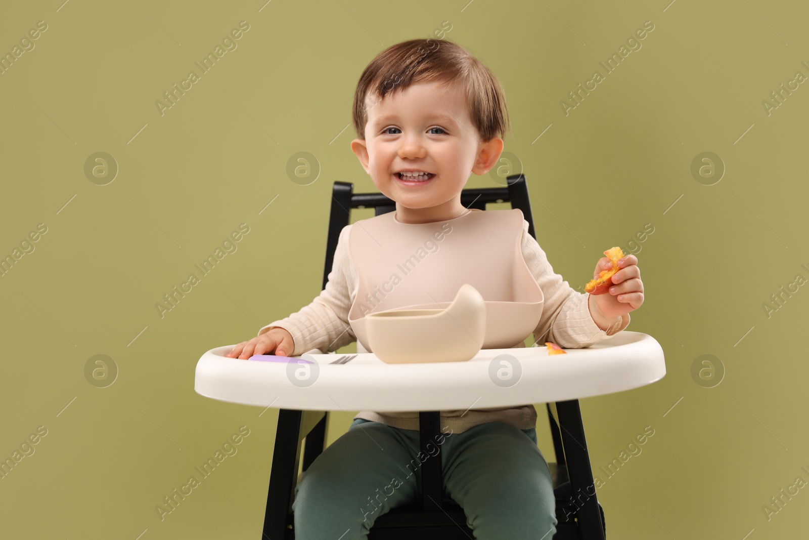 Photo of Cute little baby eating healthy food from bowl in high chair on olive background