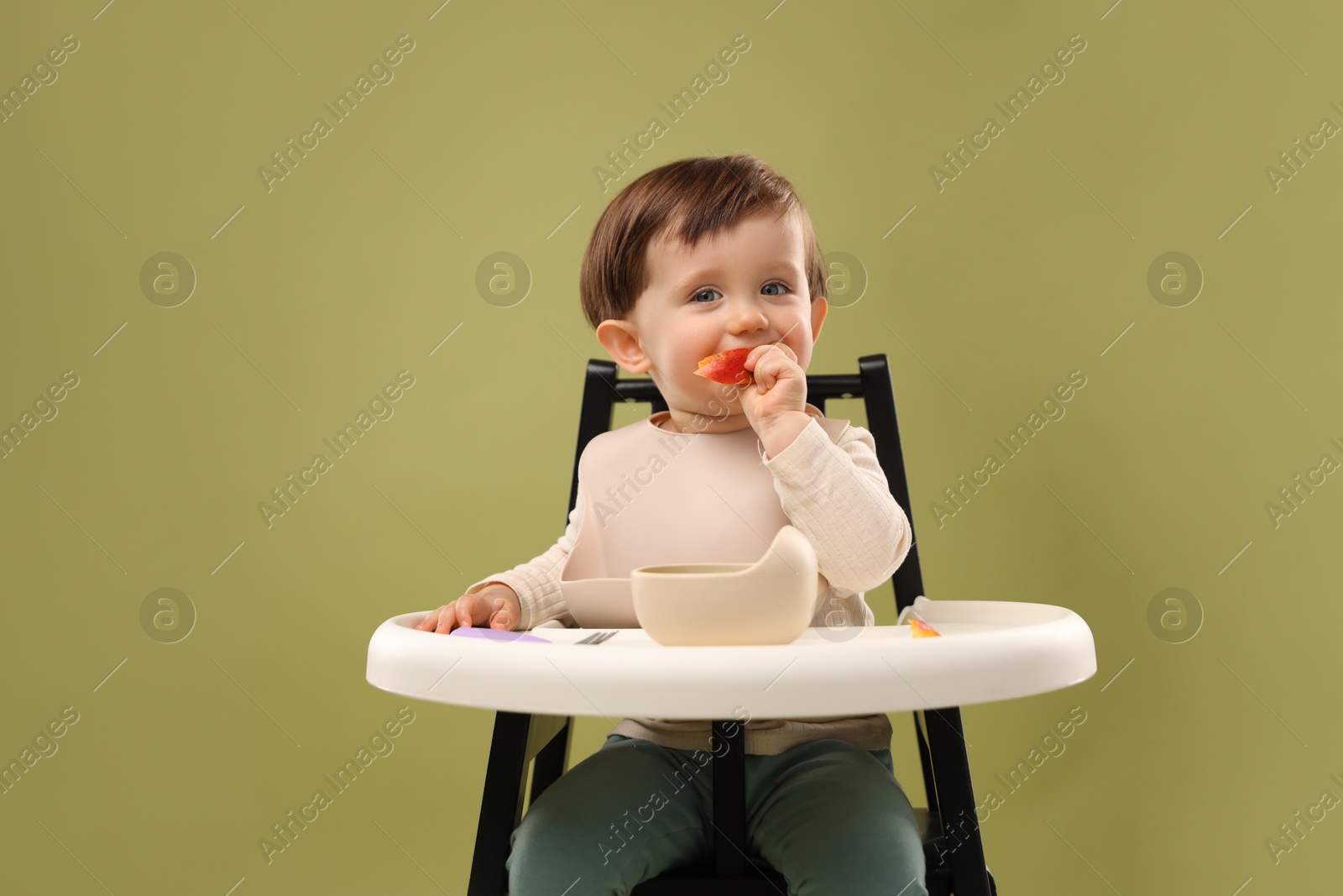 Photo of Cute little baby eating healthy food from bowl in high chair on olive background