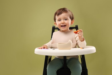 Photo of Cute little baby eating healthy food from bowl in high chair on olive background